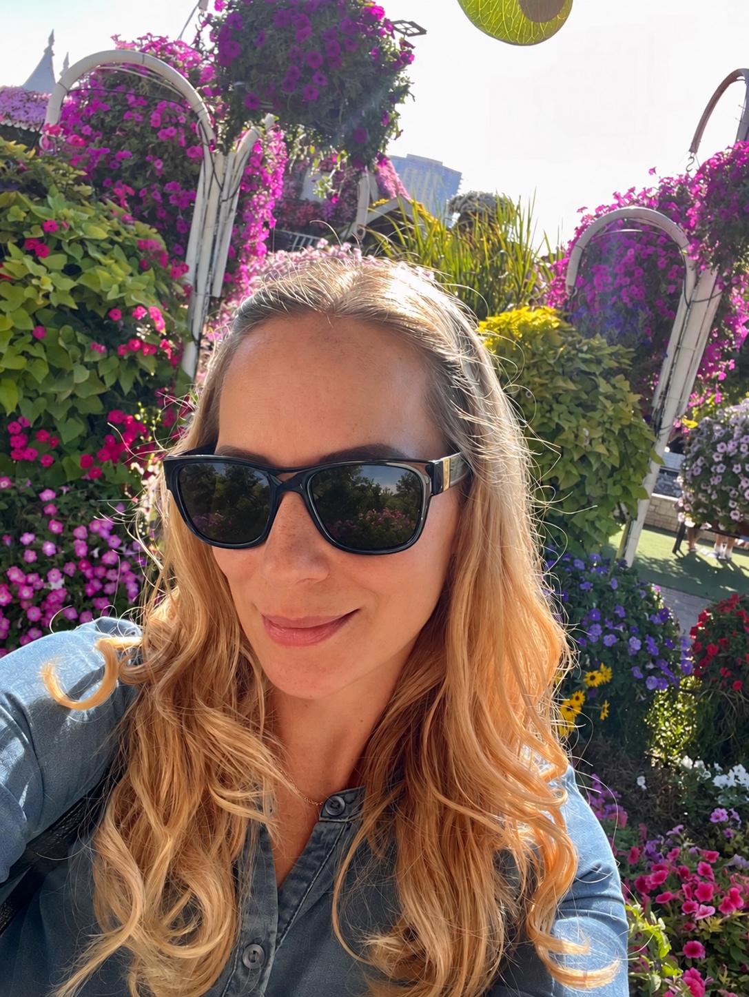 Sandra smiling in sunglasses and a denim shirt, surrounded by colourful flowers