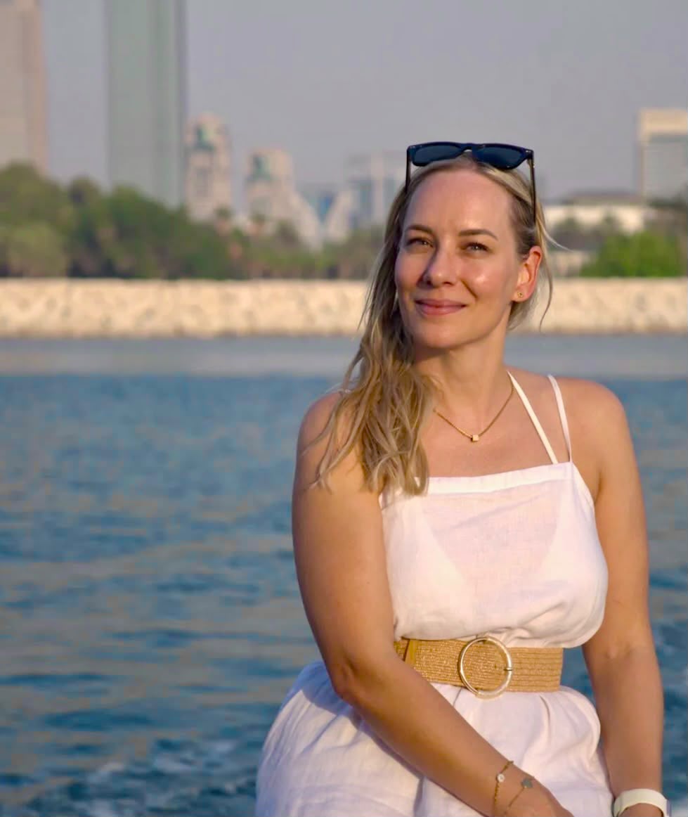 Sandra in a white dress beside the water with sunglasses resting on her head