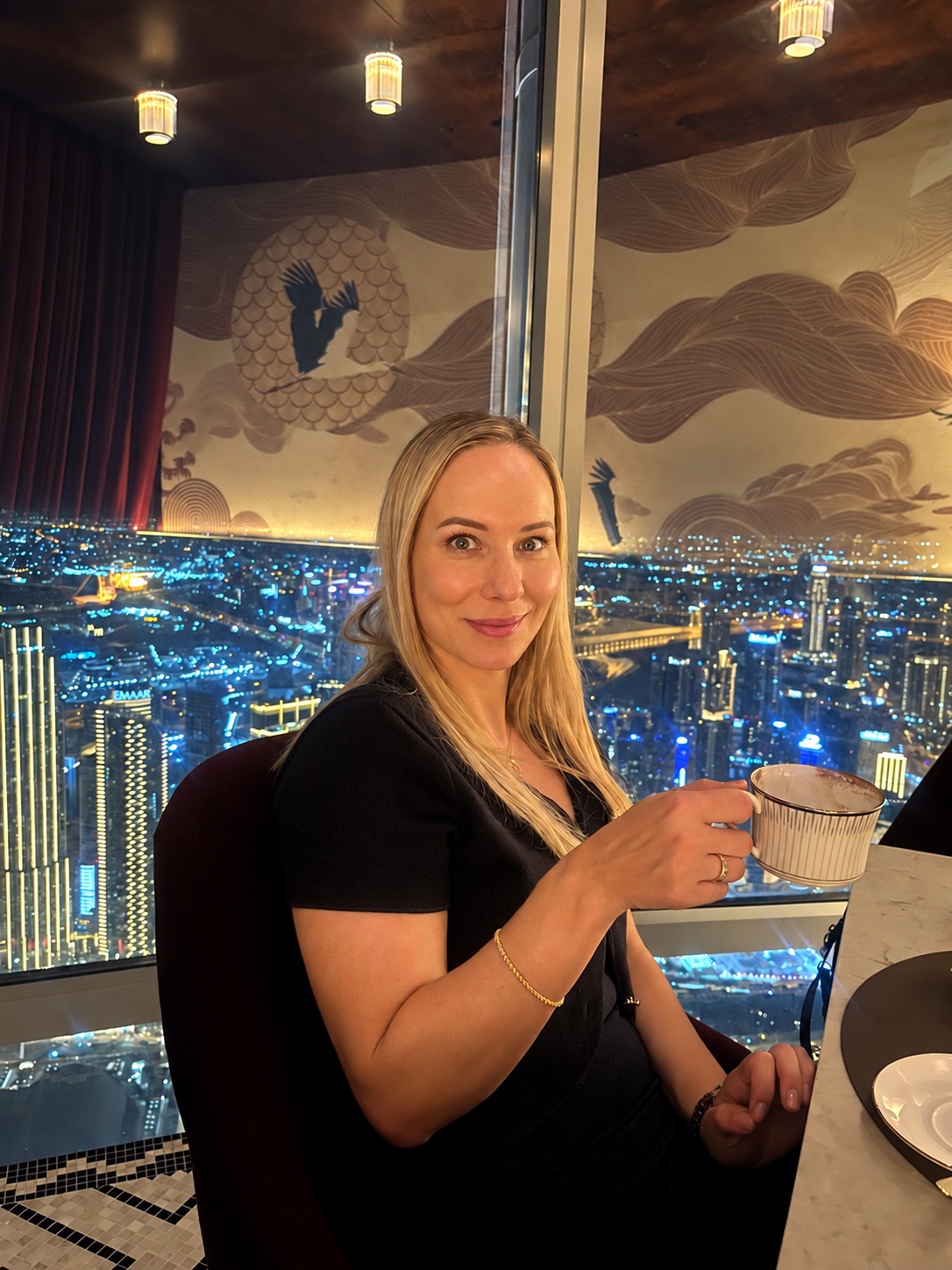 Sandra in a black dress holding a teacup beside a window overlooking city lights