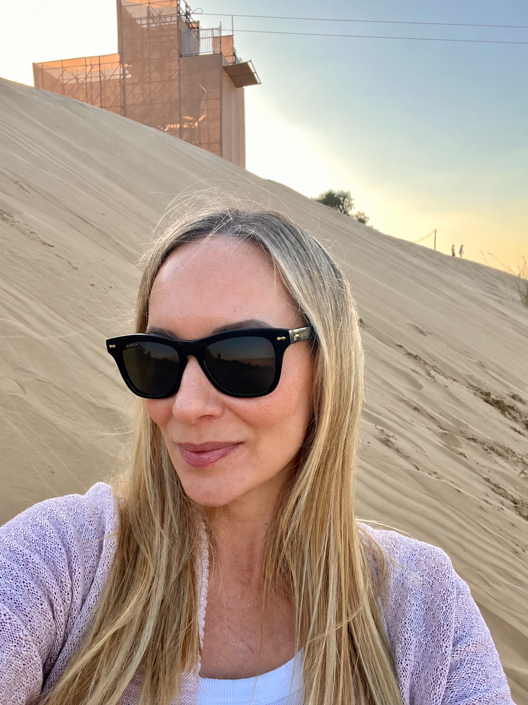 Sandra outdoors in sunglasses and a pale lilac cardigan, with sand and warm evening light behind her